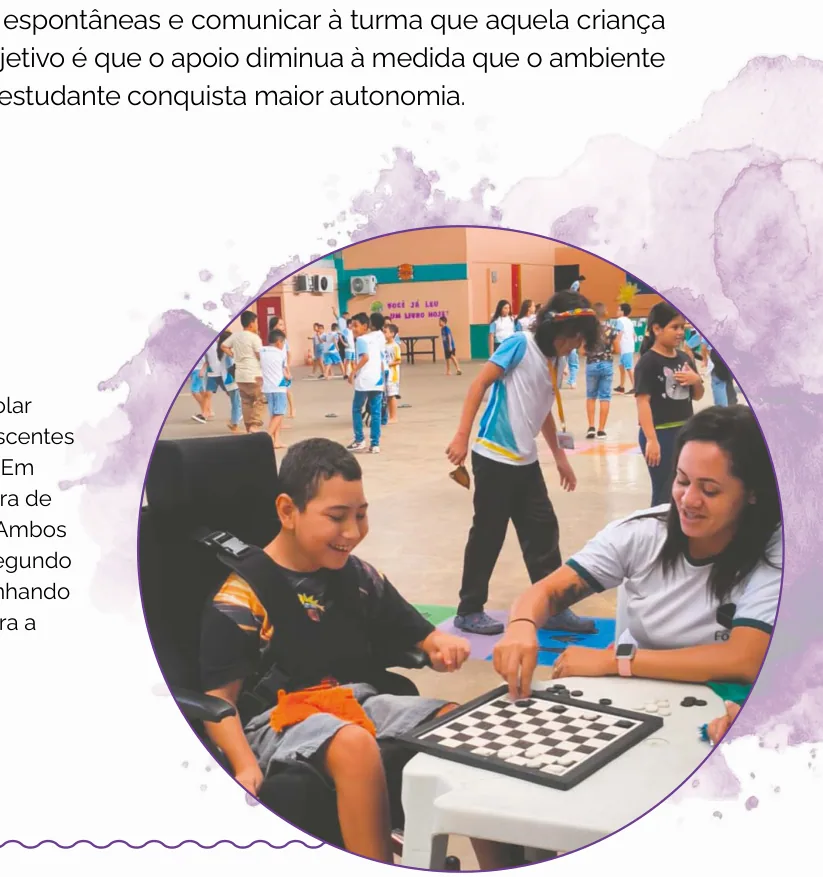 Fotografia de um menino em cadeira de rodas jogando damas com uma profissional, sorrindo, em um ginásio escolar movimentado